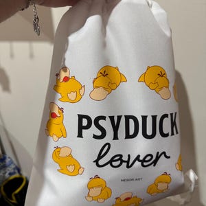 Puede incluir: Una bolsa de cordón blanca con un patrón repetitivo de personajes Psyduck amarillos. El texto PSYDUCK lover está impreso en negro en el centro, con el pequeño texto NESOR ART posicionado justo debajo de la palabra lover en la tela.