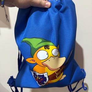 Puede incluir: Una mochila de cordón azul brillante con un estampado gráfico de una criatura amarilla con un gorro puntiagudo verde, que sostiene un pequeño escudo con un emblema oscuro. La bolsa tiene correas de cordón azules y un cierre fruncido.