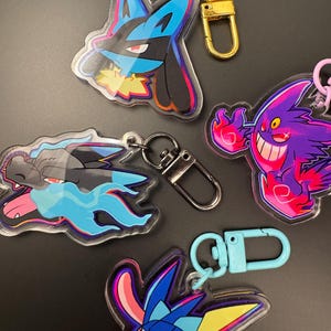 Op de afbeelding: Diverse acryl sleutelhangers met kleurrijke cartoon karakter ontwerpen. De sleutelhangers hebben metalen sluitingen in goud, zilver en blauw. De karakters zijn onder andere een blauwe en zwarte Lucario, een paarse Gengar en een blauwe en gele Greninja.