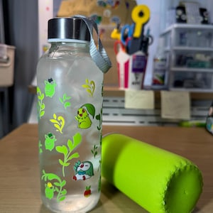 May include: A clear glass water bottle with a silver screw-top lid and a gray carrying strap. The bottle features various green and yellow cartoon character decals and leaf patterns. Beside the bottle lies a bright lime green fabric sleeve.