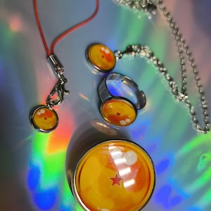 Puede incluir: Un conjunto de joyas y accesorios con un diseño amarillo y naranja y una estrella roja. El conjunto incluye un colgante, un anillo, un collar y un charm sujeto a un cordón rojo. Los artículos tienen un acabado metálico plateado.