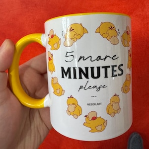 Peut inclure: Un mug en céramique blanc avec une anse et un bord jaune vif, orné d'un motif répétitif de petits canards de dessin animé jaunes dans diverses positions de sommeil. Le texte "5 more MINUTES please" est imprimé en noir, avec "NESOR.ART" en dessous.