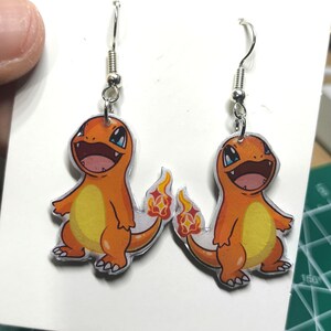 May include: A pair of orange and yellow Charmander earrings. Each earring features a smiling Charmander with a flame tail, suspended from a silver fish hook. The earrings are on a white card.