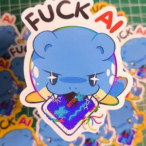 Puede incluir: Una pegatina con un personaje de dibujos animados azul y el texto "FUCK AI" en negro y rojo. El personaje sostiene una pantalla rota con la palabra "error". La pegatina tiene un borde blanco y está sobre una superficie verde.