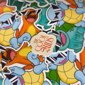 Puede incluir: Un conjunto de pegatinas coloridas con personajes de dibujos animados con gafas de sol. Las pegatinas muestran criaturas naranjas, azules y verdes con vientres amarillos. También hay una pegatina con el texto "THE COOL KIDS".