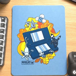 Puede incluir: Una alfombrilla de ratón rectangular de color azul claro con una consola de juegos portátil azul oscuro rodeada de ilustraciones florales amarillas y naranjas, una luna creciente y pequeñas criaturas. El texto NESOR.ART está en la esquina inferior.