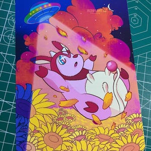 Puede incluir: Una impresión artística caprichosa que presenta una vaca de dibujos animados flotando en un cielo colorido con un OVNI. La vaca es rosa y blanca, rodeada de pétalos y girasoles. La impresión tiene el texto "NESOR ART".