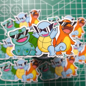 Könnte beinhalten: Eine Sammlung von Aufklebern mit Zeichentrickfiguren, darunter ein grünes Bisasam, ein blaues Schiggy und ein orangefarbenes Glumanda, die alle eine Sonnenbrille tragen. Die Aufkleber befinden sich auf einer grünen Schneidematte.