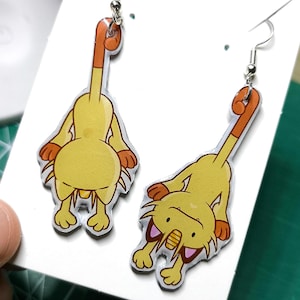 Könnte beinhalten: Ein Paar Ohrhänger mit einem gelben Cartoon-Katzen-Design und orangefarbenen Akzenten. Die Ohrringe sind an silberfarbenen Fischhaken-Ohrdrähten befestigt und werden auf einer weißen Karte präsentiert.