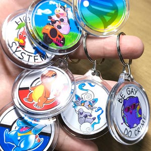 Puede incluir: Una colección de llaveros acrílicos transparentes con varios diseños de dibujos animados coloridos. Los llaveros presentan imágenes de una vaca montada en un cortacésped, un orbe de arcoíris y varios personajes de dibujos animados. Algunos llaveros tienen el texto "BE GAY DO CRIME".