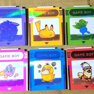 Peut inclure: Six cartes colorées de style Game Boy avec divers personnages de dessins animés. Chaque carte porte l'inscription "GAME BOY" en haut. Les personnages incluent une créature violette, Pikachu, une figurine bleue en forme de plante, Jigglypuff, Psyduck et Arcanine.