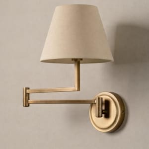 May include: Wall-mounted lamp featuring a beige fabric shade and a brass-toned adjustable arm. The lamp has a circular base and a simple design, suitable for various interior styles. The arm extends from the wall.