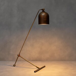 Może przedstawiać: Brązowa lampa podłogowa z kloszami w kształcie dzwonu. Lampa ma smukłą, kątową ramę i podstawę w kształcie litery T. Klosz jest umieszczony nad ramą, rzucając ciepłą poświatę. Design lampy jest minimalistyczny i nowoczesny.