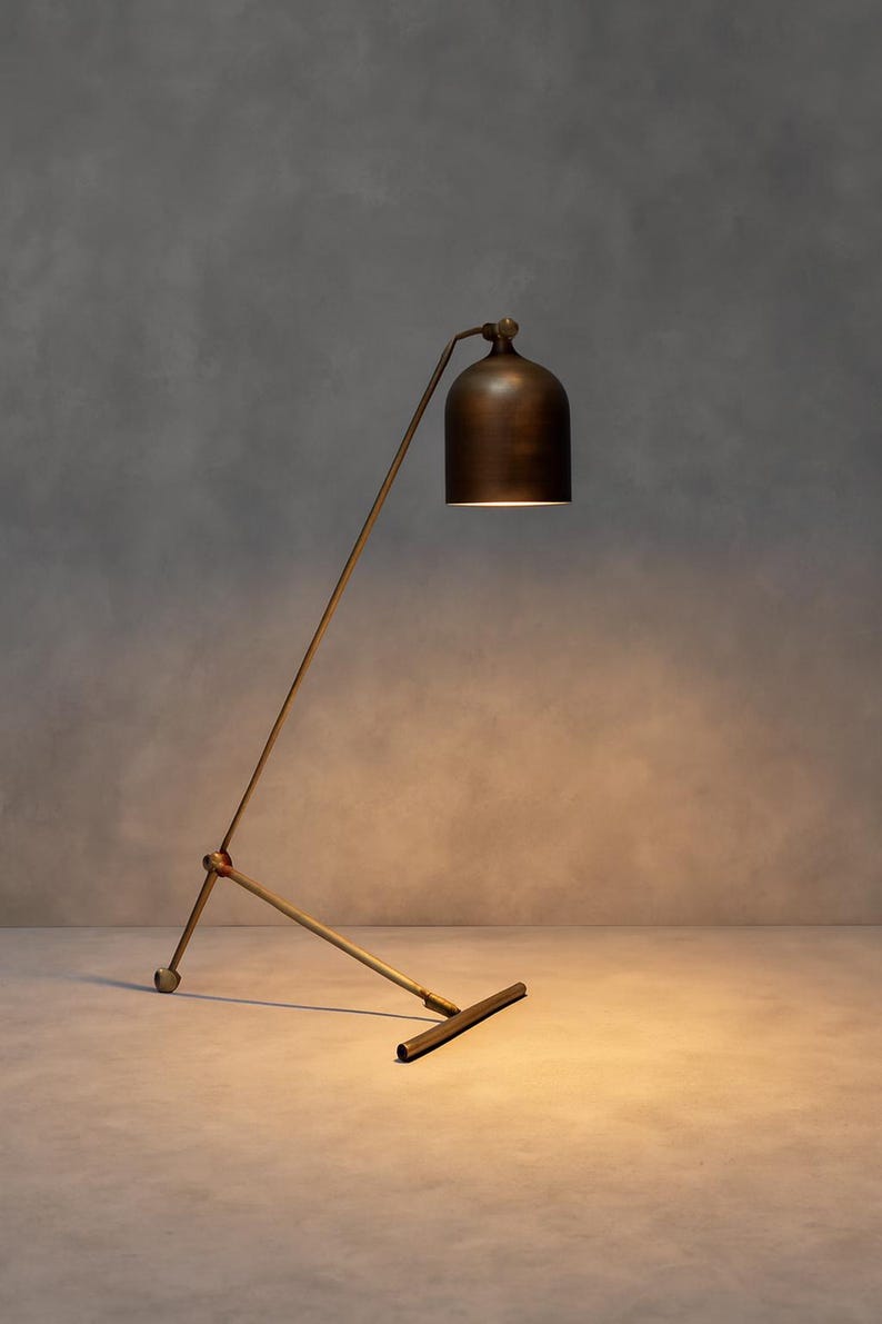 Może przedstawiać: Brązowa lampa podłogowa z kloszami w kształcie dzwonu. Lampa ma smukłą, kątową ramę i podstawę w kształcie litery T. Klosz jest umieszczony nad ramą, rzucając ciepłą poświatę. Design lampy jest minimalistyczny i nowoczesny.