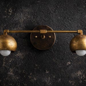 May include: A wall-mounted light fixture featuring a horizontal metal bar with a dark, weathered bronze finish. Two hemispherical shades with a textured, distressed gold-tone finish are attached to the ends, each housing a small white bulb.