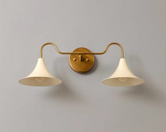 Könnte beinhalten: Eine Wandleuchte mit zwei cremeweißen, konisch geformten Schirmen. Die Leuchte hat einen gebogenen Arm in Messingfarbe und eine kreisförmige Basis. Das Design erinnert an Vintage- oder Landhausstil-Beleuchtung.