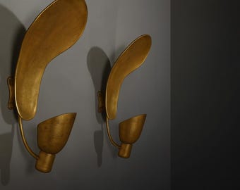 May include: Two gold-colored wall sconces with a unique, organic design. Each sconce features a curved, leaf-like element above a downward-facing, cone-shaped light fixture. The fixtures are mounted on a gray wall.
