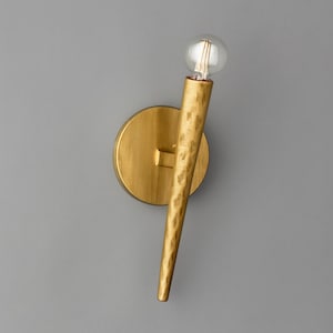 Può includere: Applique da parete color oro con un braccio allungato e strutturato e una base rotonda. L'applique presenta una lampadina rotonda e trasparente nella parte superiore. L'apparecchio è montato su una parete grigia.