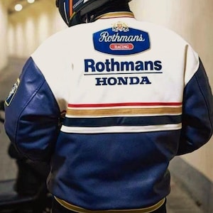 May include: A person wearing a blue, white, and gold Shoei helmet and a white and blue Rothmans Honda racing jacket. The jacket has the Rothmans Racing logo on the back and features red and gold stripes.