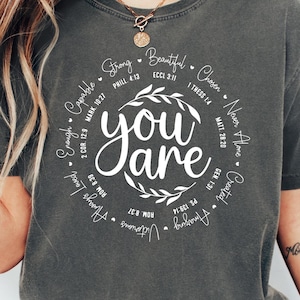 May include: Dark grey t-shirt with the words "You Are" in a white, circular design. The design includes inspirational words and phrases. The model is wearing blue jeans and white trainers.