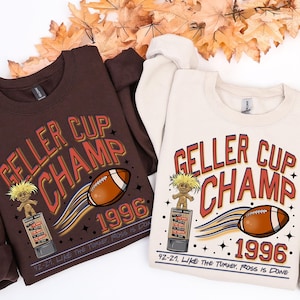 May include: Two sweatshirts, one brown and one cream, feature the text "GELLER CUP CHAMP 1996" with a football graphic and a troll doll. The text "92-21, LIKE THE TURKEY, ROSS IS DONE" is also printed on the sweatshirts.