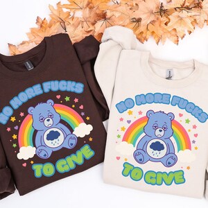 May include: Two sweatshirts, one brown and one cream, each featuring a blue bear graphic with a rainbow and the text "NO MORE FUCKS TO GIVE." The bear has a sad expression and a rain cloud on its belly. The sweatshirts are displayed on a white surface with autumn leaves.