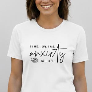 May include: White t-shirt with the text "I came. I saw. I had anxiety. So I left" in black script. The word "anxiety" is in a stylized font with a small graphic. The shirt is worn by a person.