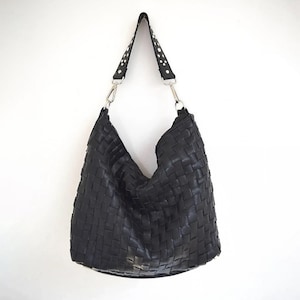 May include: Black woven leather shoulder bag with a studded strap and silver-tone hardware. The bag has a large, open design and a single shoulder strap. The bag is hanging against a white background.