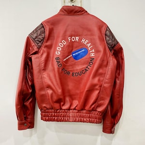 Könnte beinhalten: Rote Lederjacke mit Rundkragen und geripptem Saum. Auf dem Rücken befindet sich eine Grafik einer blau-weißen Pille mit dem Text "GOOD FOR HEALTH BAD FOR EDUCATION". Die Schultern haben ein gestepptes braunes Design.