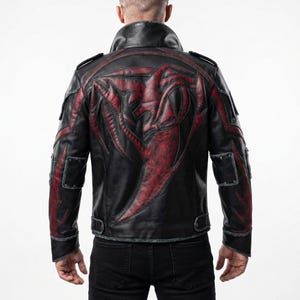 Könnte beinhalten: Eine schwarze Lederjacke mit einer großen, erhabenen rot-schwarzen Grafik im Tribal-Stil auf dem Rücken. Die Jacke verfügt über einen hohen Kragen, Schulterklappen und dekorative Einsätze an den Ärmeln mit silberfarbenen Metallnieten.