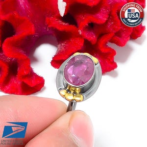 Peut inclure: Une bague en argent orn&eacute;e d'une grosse pierre rose ovale, avec des d&eacute;tails dor&eacute;s. La bague est tenue dans une main devant un fond de fleur rouge vif. Le design de la bague est &eacute;l&eacute;gant et accrocheur.