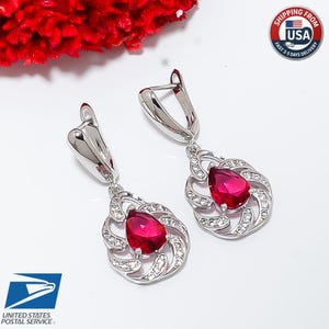 May include: A pair of silver-toned drop earrings featuring a central pear-shaped red gemstone surrounded by a swirling frame of small clear stones. The earrings have a secure hinged clasp mechanism and a polished metallic finish.