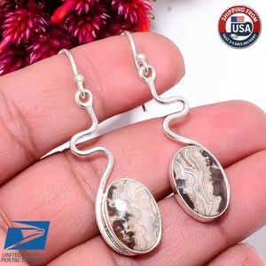 May include: A pair of silver dangle earrings featuring oval-shaped stones with intricate, wavy, cream and dark brown banded patterns. The earrings have a distinctive, curved, serpentine silver wire design connecting the hook to the stone setting.