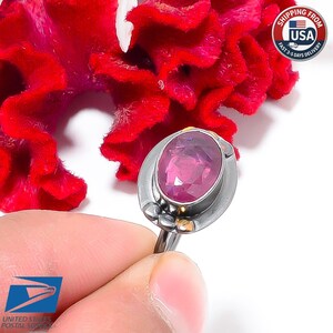May include: A silver ring with an oval, pink gemstone. The ring has a unique design with small gold accents. The background features a vibrant red flower and a shipping label.