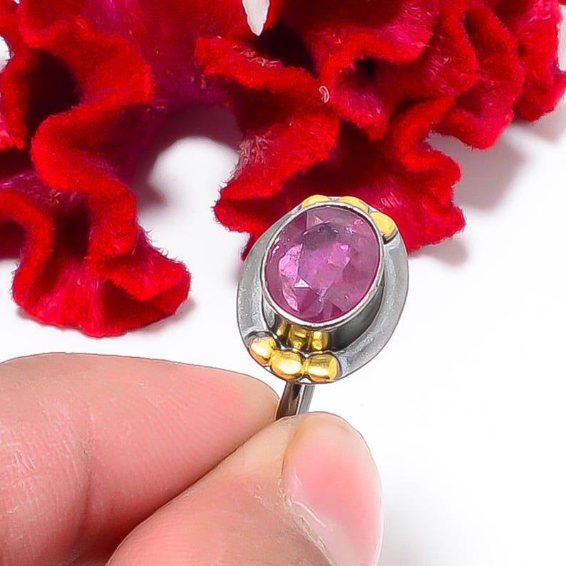 Peut inclure: Une bague en argent orn&eacute;e d'une grosse pierre rose ovale. La bague pr&eacute;sente des d&eacute;tails dor&eacute;s autour de la pierre et &agrave; la base. La bague est tenue dans une main, avec une fleur rouge en arri&egrave;re-plan.