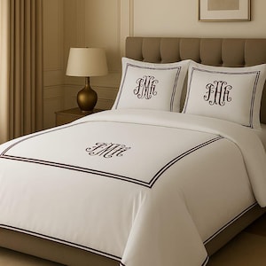 May include: White bedding set with a monogram design in a dark brown colour. The set includes a duvet cover and two pillowcases, all with a border and monogram. The bed is made up with a neutral colour scheme.
