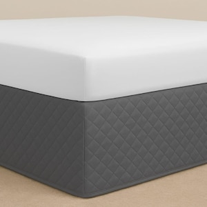 May include: A bed with a white fitted sheet and a dark grey bed skirt. The bed skirt has a diamond-quilted pattern. The bed is on a light tan surface.