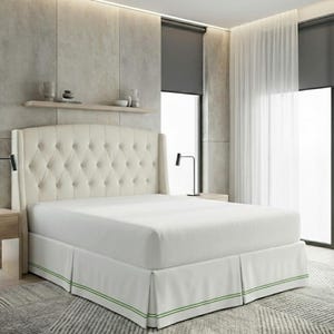 May include: A cream-coloured upholstered bed frame featuring a high, tufted headboard with button detailing and side wings. The bed is dressed in white bedding and a white bed skirt accented with two thin, horizontal green stripes along the bottom.