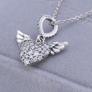 May include: A silver necklace featuring a heart-shaped pendant with small, clear stones and attached wings. The pendant is suspended from a delicate chain. The necklace has a heart-shaped bail with clear stones and the text "S925".