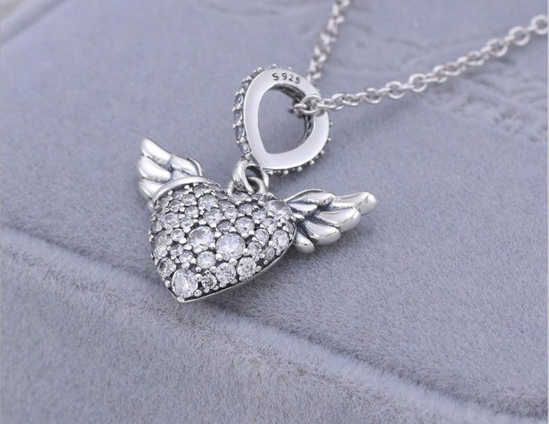 May include: A silver necklace featuring a heart-shaped pendant with small, clear stones and attached wings. The pendant is suspended from a delicate chain. The necklace has a heart-shaped bail with clear stones and the text "S925".