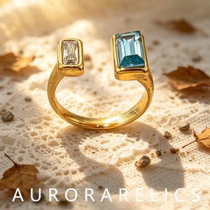May include: An open-style gold ring featuring two rectangular stones: a smaller clear white baguette-cut stone and a larger light blue emerald-cut stone. The gold band has a textured surface, and the text AURORARELICS is visible at the bottom.