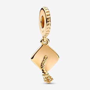 May include: A gold-colored charm featuring a graduation cap design. The charm has a square cap with a tassel hanging from the corner. The top of the charm has a ring for attaching to a bracelet or necklace.
