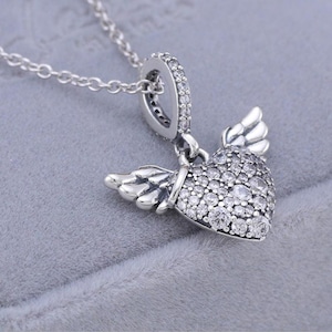 May include: A silver necklace featuring a heart pendant with wings. The heart is encrusted with small, clear gemstones. The wings are detailed with feather-like designs. The necklace chain is delicate and silver.