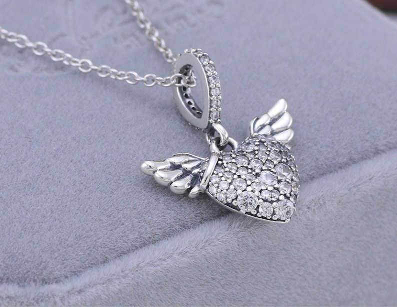 May include: A silver necklace featuring a heart pendant with wings. The heart is encrusted with small, clear gemstones. The wings are detailed with feather-like designs. The necklace chain is delicate and silver.