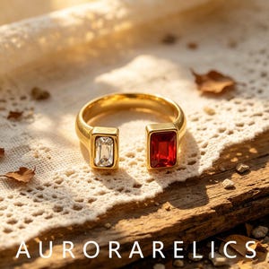 May include: A gold-toned open ring featuring two rectangular stones, one clear and one deep red, set at each end of the band. The text AURORARELICS is displayed at the bottom of the image. The ring has a polished finish and a simple design.