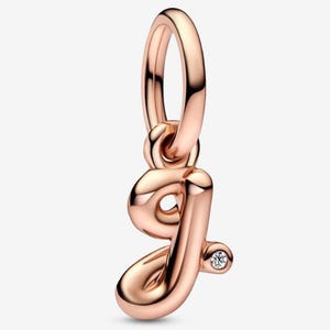 May include: Rose gold charm in the shape of the letter "J" with a small, clear gemstone. The charm has a polished finish and a loop at the top for attaching to a bracelet or necklace. The charm is a jewelry item.