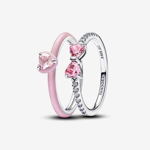 May include: Two rings. One is a light pink band with a heart-shaped pink gemstone. The other is a silver band with a row of small clear gemstones and two heart-shaped pink gemstones. The word "PANDORA" is engraved on the silver band.