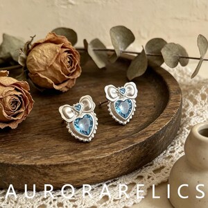 May include: A pair of silver-toned stud earrings featuring a heart-shaped light blue crystal set within a scalloped border, topped with a decorative bow and a tiny matching blue crystal accent, designed with a post back for secure attachment.