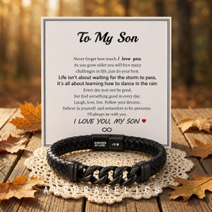 May include: A black braided leather bracelet with a matte black metal chain link centre section. The clasp features the text SHIELDED BY LOVE and J.B. with a heart symbol. It is displayed in front of a white card with the printed text To My Son.