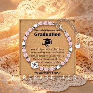 May include: A bracelet featuring light pink beads, silver-toned spacers, and a round charm with a compass design. The bracelet is displayed on a rectangular card with the text Congratulations on your Graduation, a graduation cap icon, and an inspirational message about new beginnings and the future.